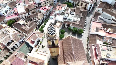 aerial drone shot of historic church with patterned tower surrounded by narrow streets, whitewashed houses, tiled rooftops, and small plaza with trees in old town area of ronda, spain