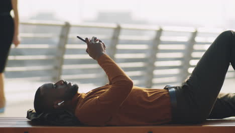 hombre afroamericano acostado en un banco y usando un teléfono inteligente