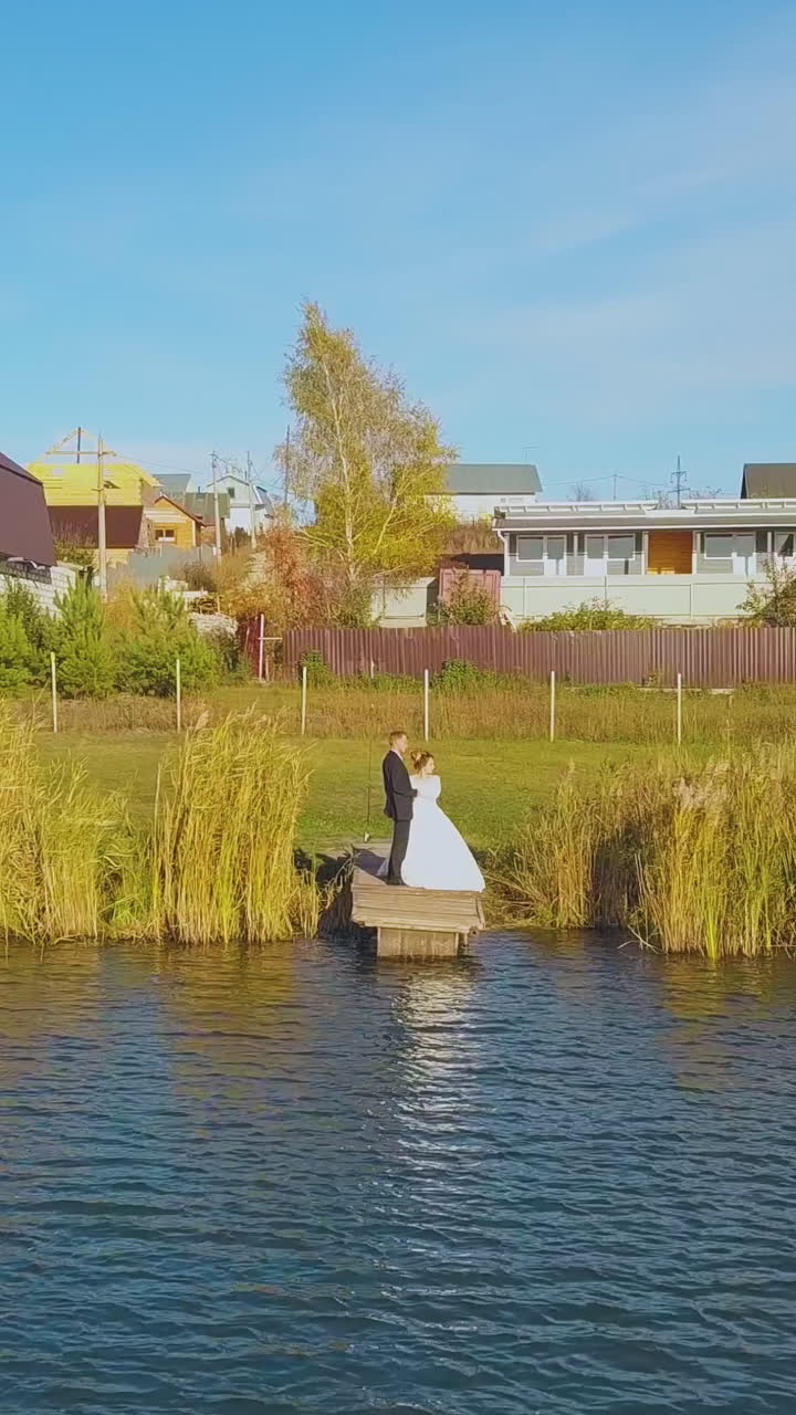 young groom and bride stand on wooden pier near calm river at cottage houses on sunny day aerial view