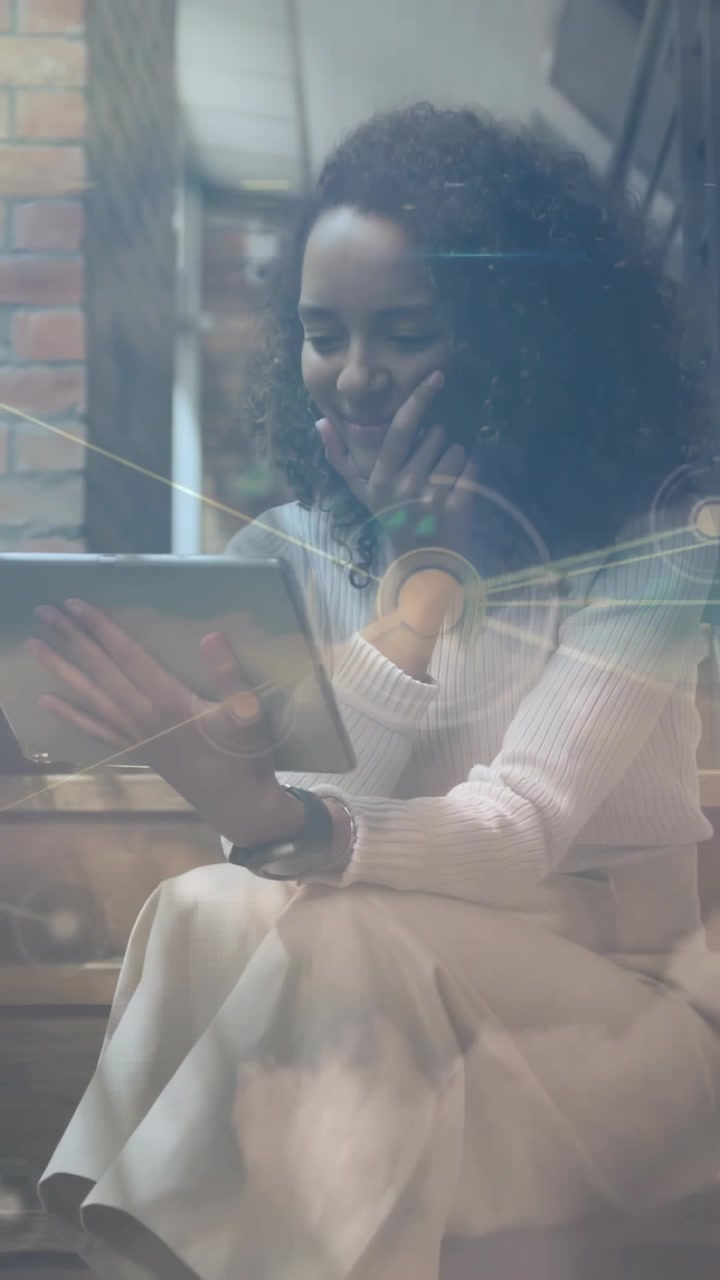 Adult woman sitting by window using tablet reading and swiping, causing tech overlay, smiling