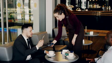 cheerful caucasian businessman sitting with his african american partner and paying online bill using his smartphone in modern cafe. young waitress using cash terminal.