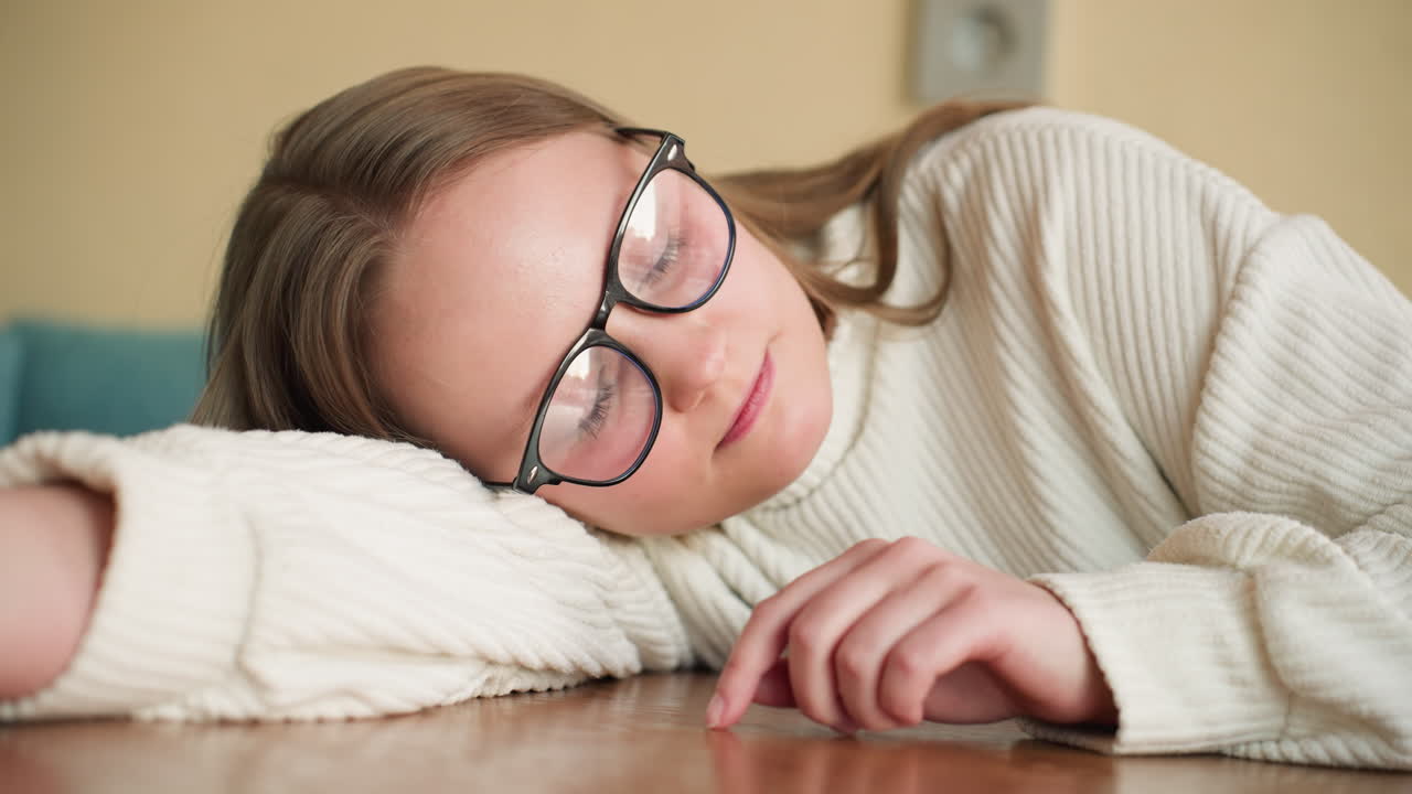 Young woman in glasses rests head on cozy white sleeve while smiling softly and gently tracing finger on wooden table, expressing calm creative thoughtfulness