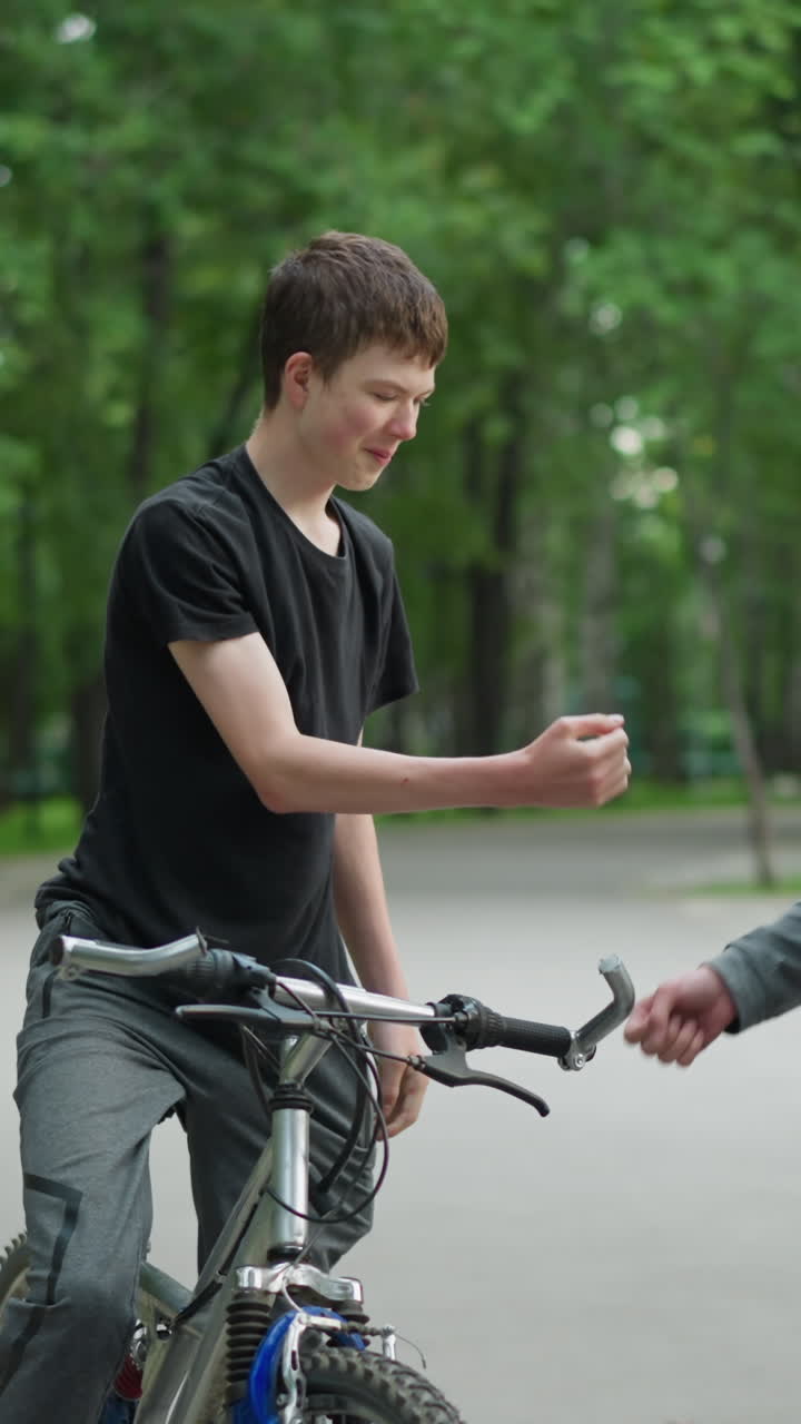 dos hermanos en bicicletas comparten un momento amistoso en un entorno de parque, un niño en una camiseta gris espera mientras el otro cabalga para saludarlo con un golpe de nudillo, el fondo cuenta con árboles verdes exuberantes