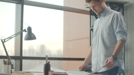 bearded man (hipster) standing faces to panoramic window and looks at blueprints. young architect in stylish loft office works with documents. creative architect thinking on the big drawings.