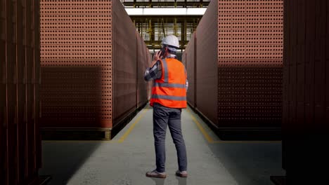 full body back view of asian male engineer with safety helmet talking on smartphone while standing with red brick packed in stacks are stored