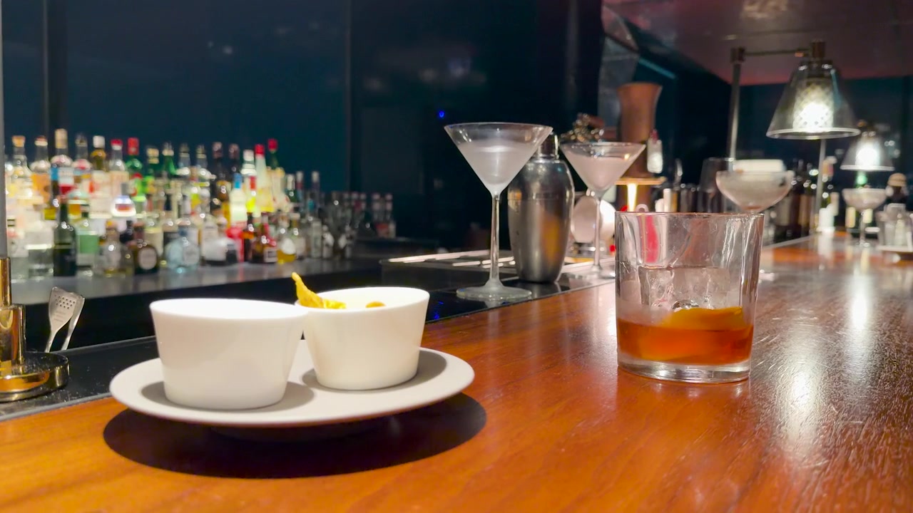 Một ly cocktail whisky đặt trên quầy bar gỗ cùng với đồ ăn nhẹ trong một phòng chờ sang trọng.