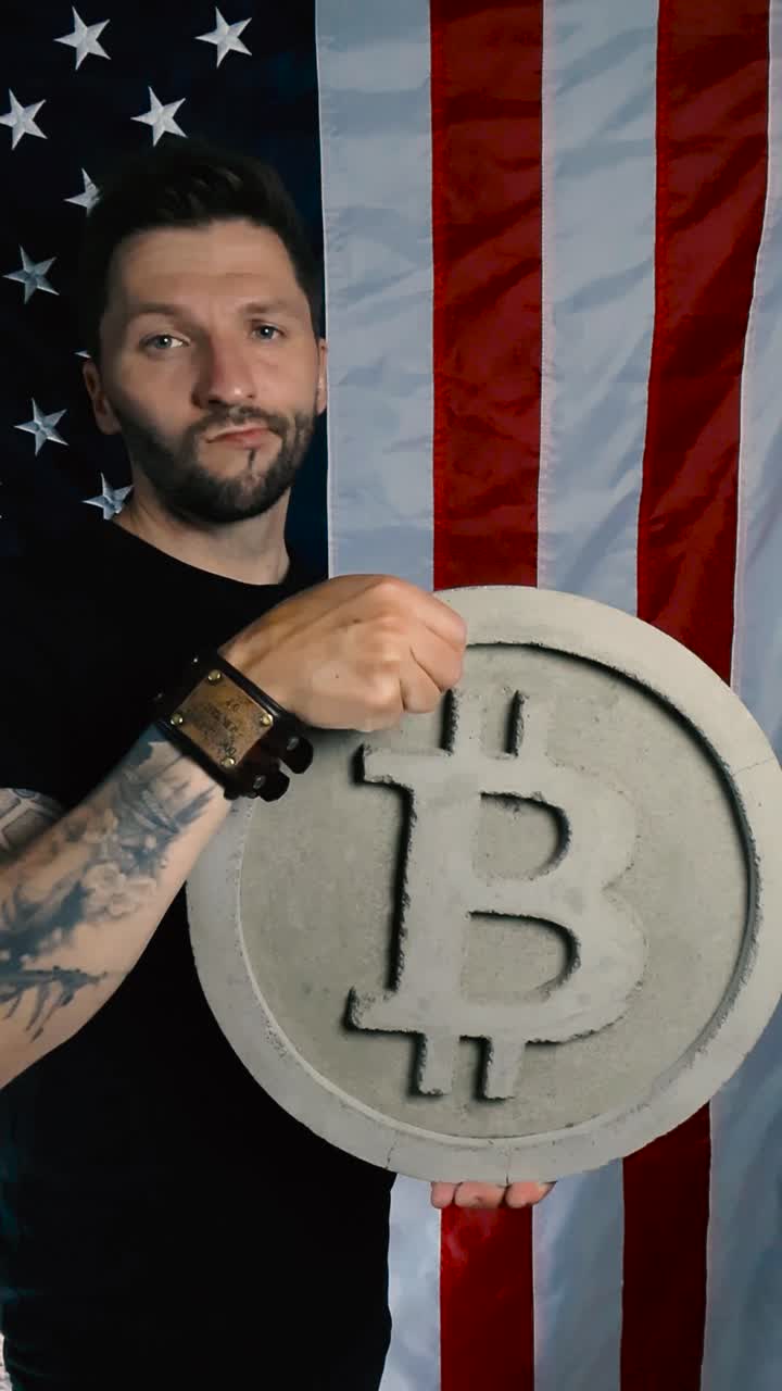 Tattooed white man lifting up a large Bitcoin cryptocurrency money gray colored sculpture and begins to shake his head while not happy in front of a vertically hanging US American flag, slow motion