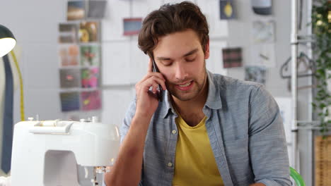 Male-Owner-Of-Fashion-Business-Making-Call-On-Mobile-Phone-At-Sewing-Machine-In-Studio