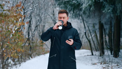 Man wearing black jacket stands in the winter park. Caucasian male drinks coffee and looks at his smartphone.