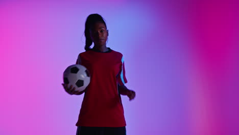 Studio-Portrait-Of-Female-Football-Or-Soccer-Player-Wearing-Team-Shirt-Holding-Ball-Under-Her-Arm-Shot-Low-Key-Against-Colourful-Mixed-Lighting-2