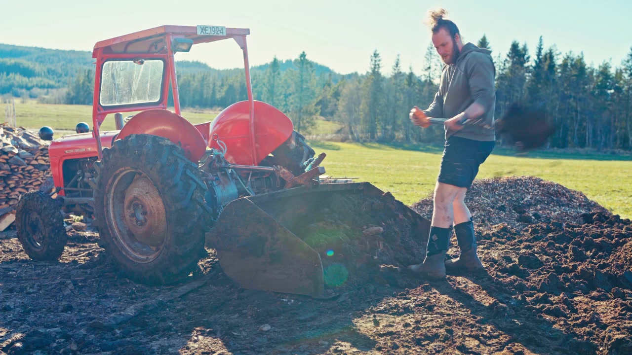 Person Using Shovel On Muddy Ground Next To An Agricultural Tractor In A Rural Background. Static Shot