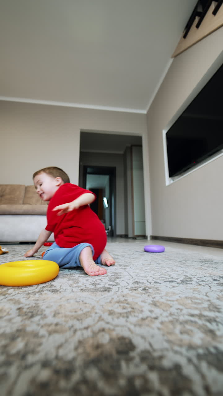 Cute baby boy sitting on the floor takes off his sock. Barefoot toddler plays at home cheerfully. Low angle view. Vertical video.