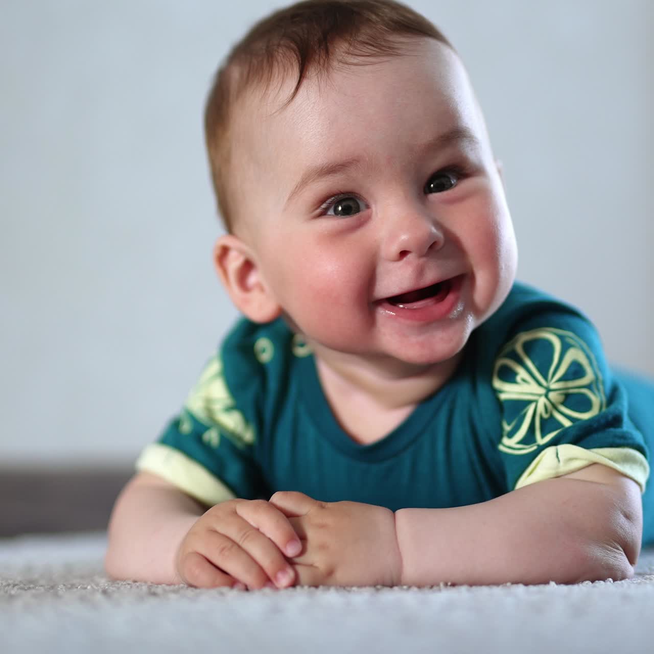 Adorable smiling kid lies on the floor looking at camera. Cheerful kid banging hands by the floor actively. Close up