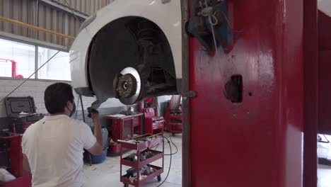 Dirty-latin-male-mechanic-removing-the-fender-liner-of-a-white-car-without-wheel-at-a-workshop-station-in-mexico-latin-america