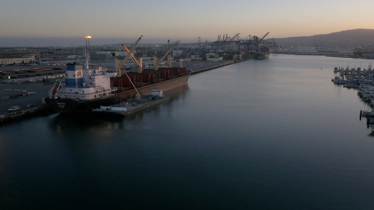 A cargo ship docked at an industrial port with cranes at dusk