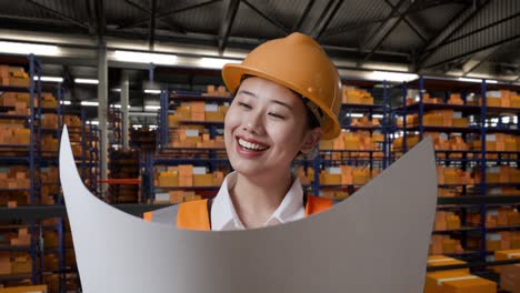 close up de ingeniera asiática con casco de seguridad mirando el plano en sus manos y mirando a su alrededor mientras estaba de pie en el almacén con estantes llenos de bienes de entrega