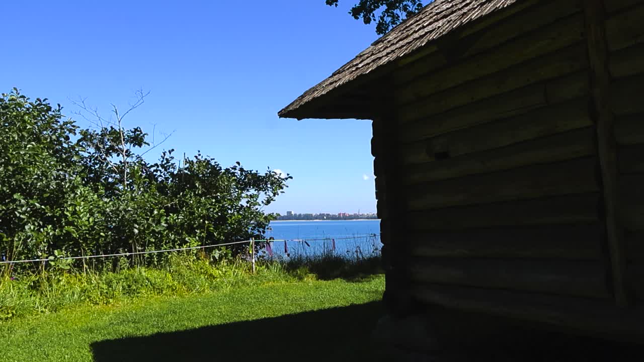 Old traditional and vintage log cabin with a splinter roof or wood chip roofing at the baltic sea blue ocean seaside shoreline beach during a summer sunny day, horison visible besides the trees