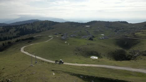 Tractor-on-dirt-road-driving-in-highland-mountain-plateau,-Velika-Planina,-village-in-background