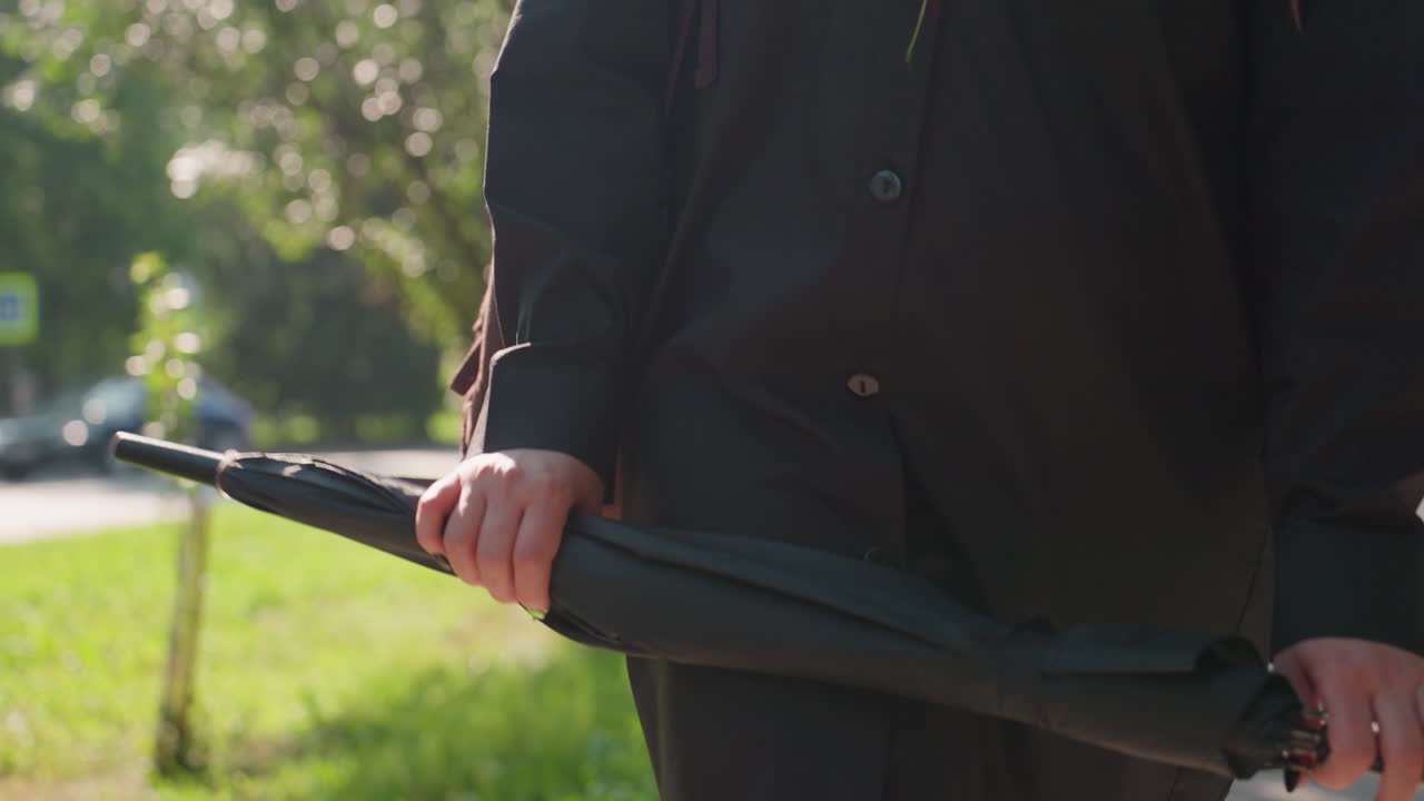 공원에서 우산을 든 손, 검은 코트를 입은 슬픈 여자가 햇빛이 비치는 길가에서 잠시 멈춰 서 있습니다. 빨간 매니큐어를 칠한 손톱과 숄더백이 보이고, 조용한 사색과 생각에 잠긴 분위기, 은은한 햇빛과 부드러움이 느껴져요.