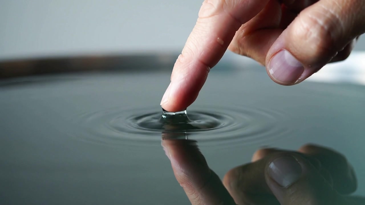 Abbassando la punta del dito indice destro che tocca l'acqua in una ciotola bassa sul tavolo, formando una goccia e increspature.