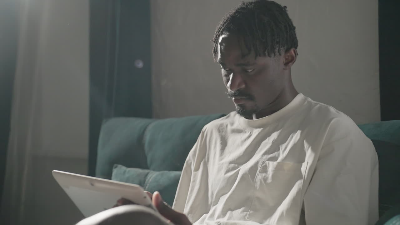 Champ seated on teal sofa, focused on tablet with natural light streaming from the window. Peaceful, thoughtful moment in a calm, cozy living room with modern, minimalist decor