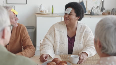 waist-up over shoulder of cheerful older people sitting at table, playing forehead detective game, caucasian man asking questions of his character, answering, african woman smiling, applauding
