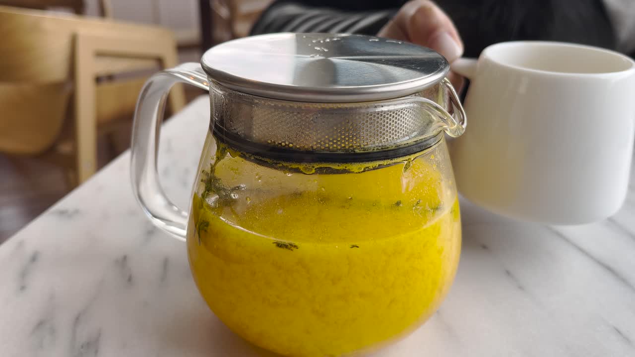 Closeup of glass teapot filled with vibrant yellow turmeric herbal tea, brewed with fresh herbs, on marble table, beside cup held by person.