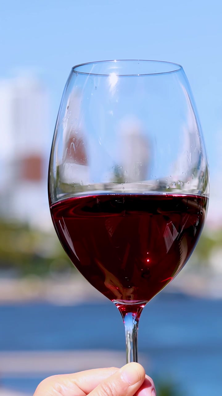 Una mano agita delicatamente un bicchiere di vino rosso contro uno sfondo urbano luminoso e all'aperto. La luce naturale del giorno evidenzia il movimento e i riflessi del vino