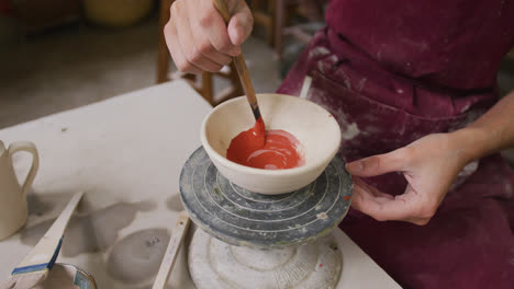 mid section of female potter wearing apron painting pot on potters wheel at pottery studio