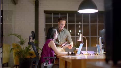 colegas masculinos y femeninos enfocados hablando y usando portátiles y tabletas por la noche en la oficina