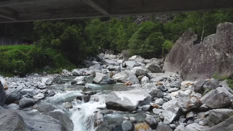 Old-bridge-over-rocky-mountain-river-in-Switzerland,-aerial-low-angle-view