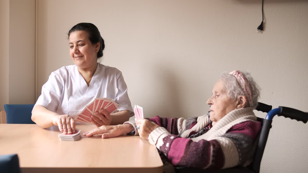 Smiling nurse playing cards with old woman using wheelchair