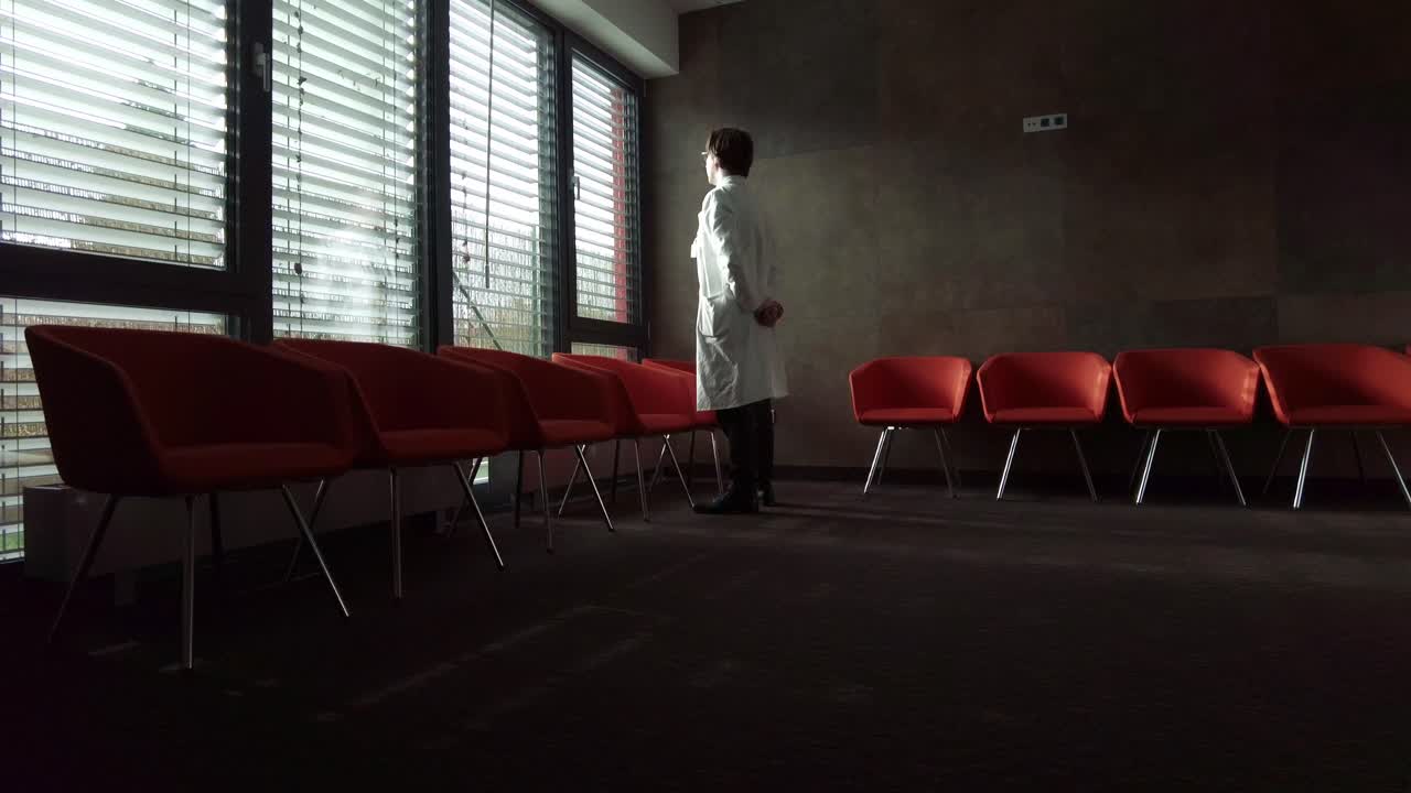 A young doctor with a white coat looking and thinking at the window with the sun's rays, taking a break between services in a dark room with chairs