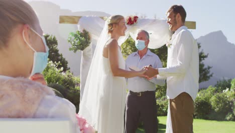 caucasian bride groom and wedding officiant wearing face mask standing at outdoor altar