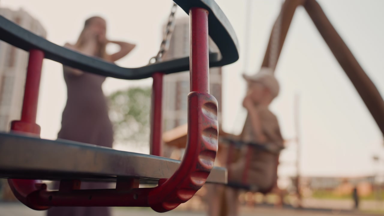 Una donna e un bambino interagiscono in un parco giochi all'aperto, con un focus sui dettagli di un'altalena rossa. Lo sfondo mostra edifici urbani sotto una luce diurna tenue mentre si dedicano ad attività di gioco casuali.