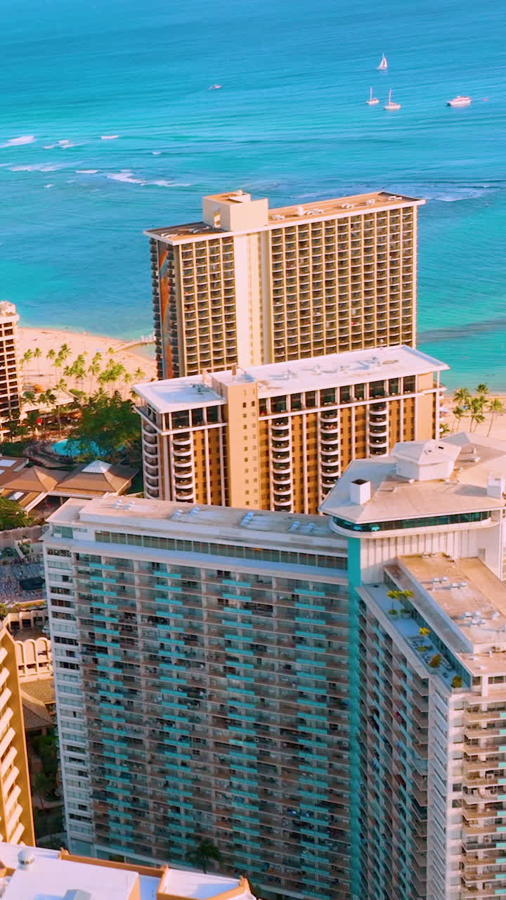 Edifici resort a più piani vicino alla costa con acqua oceanica turchese e piccole barche sullo sfondo. Veduta aerea di hotel di lusso e dell'oceano a Honolulu. Video verticale.