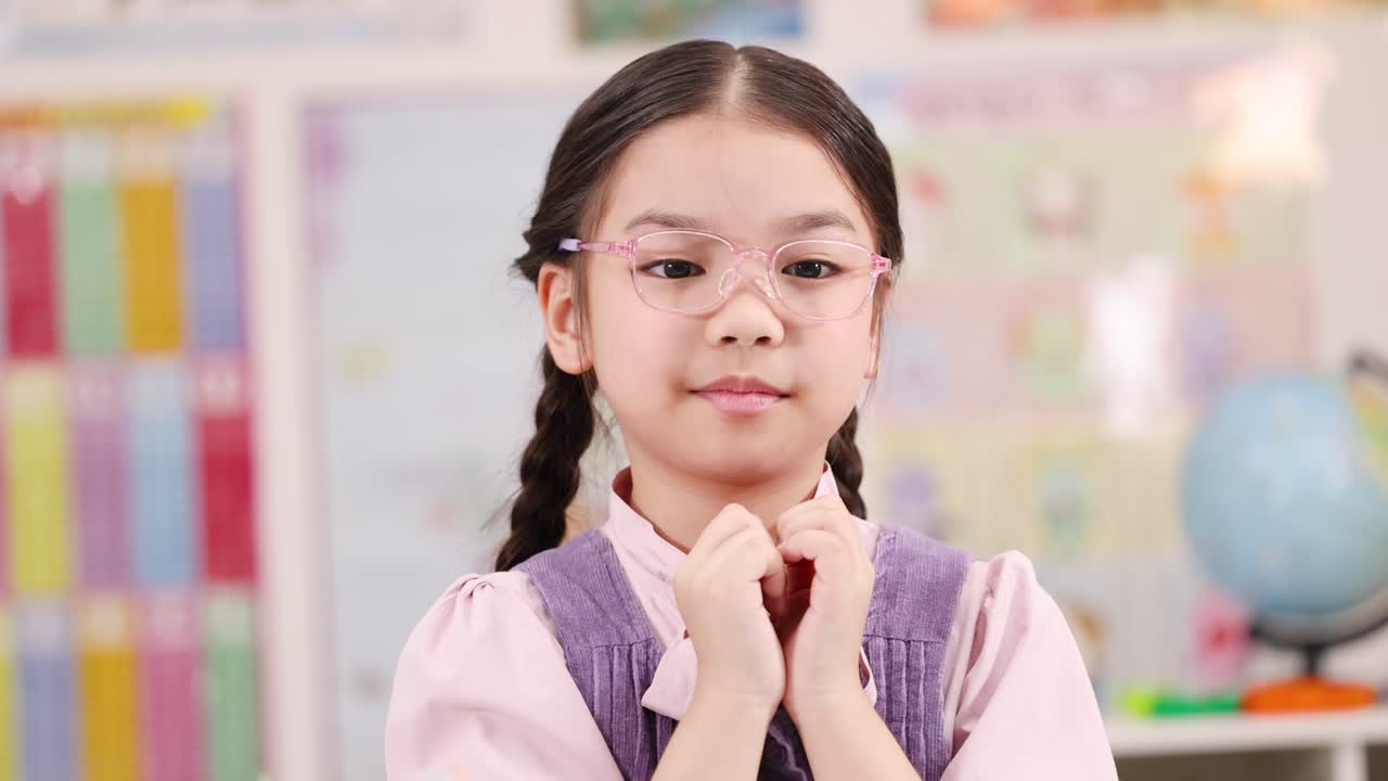 Young girl with glasses smiles gently, hands clasped, in bright, cheerful classroom environment