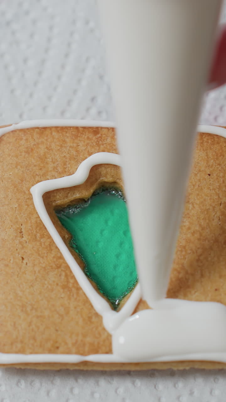 Close-up of person icing a cookie with white frosting, creating intricate designs around a green filling, on textured paper, showcasing the baking and decoration process