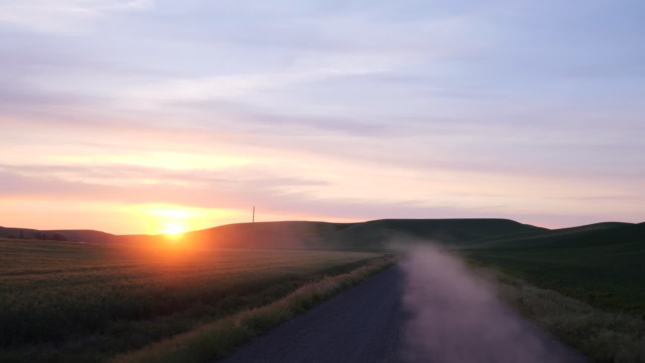 Aerial footage captures a tranquil dusty road winding through expansive golden fields at sunset. The serene countryside setting, bathed in warm light, offers a captivating sky perspective