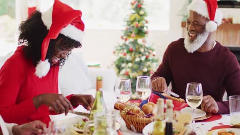 familia afroamericana en sombreros de santa hablando y sonriendo mientras están sentados en la mesa de comedor almorzando