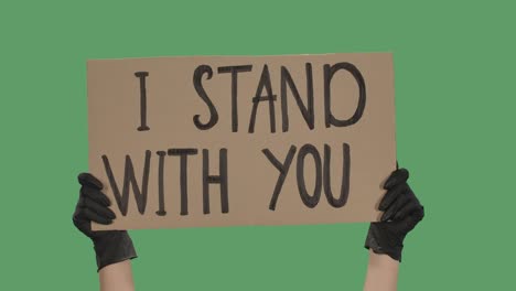 the hands of an unknown person in black gloves raise a cardboard poster i stand with you. equality and unity concept. isolated a green screen, chroma key. close up. slow motion