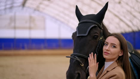 mid-aged young woman with long dark hair holding a horse by the bridle. close up. woman caressing the animal before riding it.