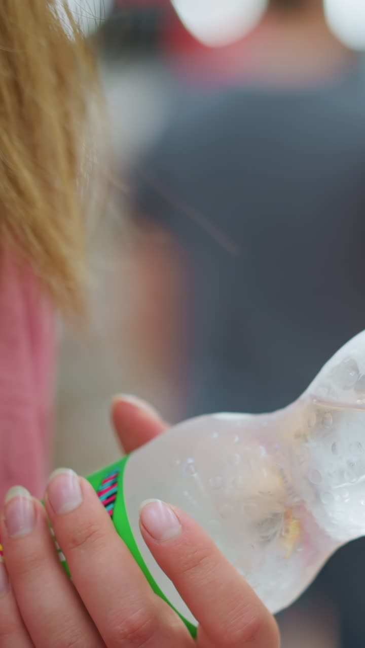 한 여인이 다른 사람의 뒤에 있는 쇼핑몰에서 차가운 병병 물을 들고 있으며, 잘 조명된, 흐릿한 배경에는 다른 사람들이 돌아다니고 있으며, 병은 고 차가워서 습기가 있습니다.