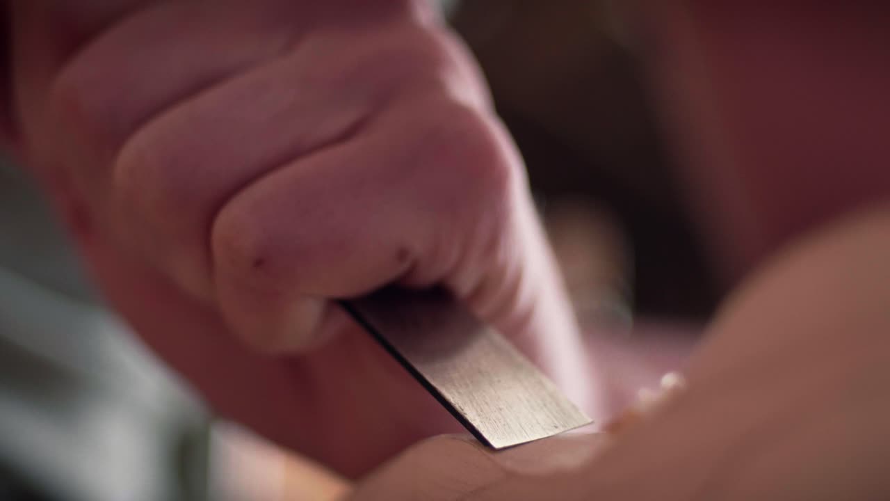 Female artisan craftsman's hands carefully using a sharp metal chisel to shape a piece of light-colored wood in a carpentry workshop. Slow motion