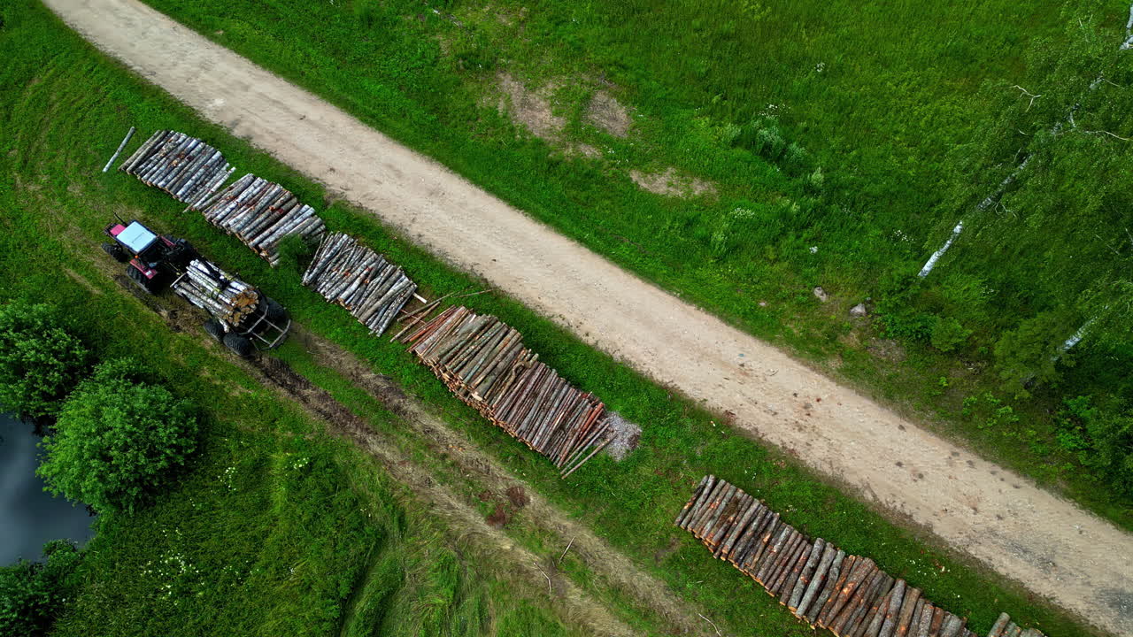 A tractor beside piles of timber along a rural dirt road through green fields, aerial view