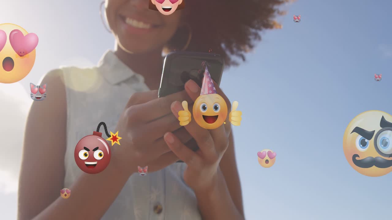 animación de emoticones sobre la sección media de una mujer biracial que usa un teléfono inteligente al aire libre