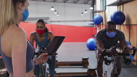 Entrenadora-De-Fitness-Femenina-Caucásica-Con-Mascarilla-En-El-Gimnasio-Sosteniendo-Una-Tableta