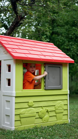 charming boy opens the window of the play house from inside. kid leaves the house and walks by the grass. vertical video.