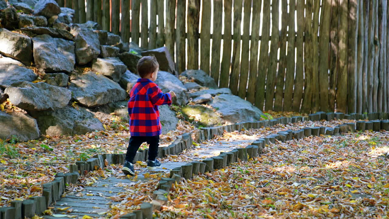 Kid in red and blue shirt runs by the narrow path along the pile of rocks. Toddler boy approaches the wooden poles fence.