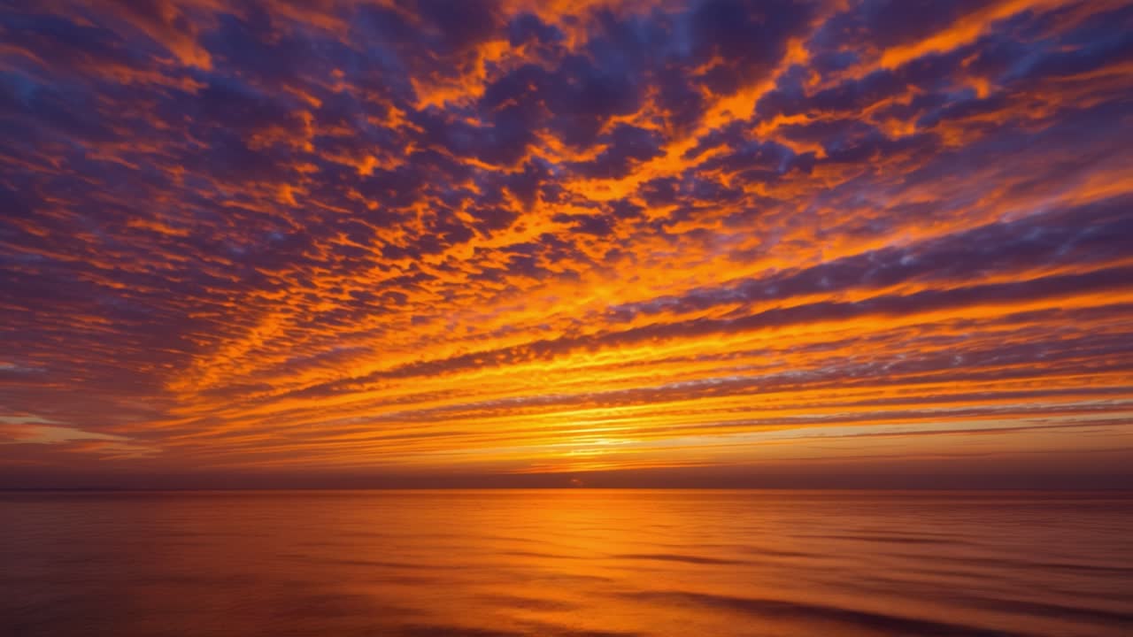 A breathtaking view unfolds as the sun sets over the tranquil ocean. The sky is filled with dramatic clouds painted in shades of orange, pink, and purple, creating a stunning contrast against the deep blue water below. Gentle waves ripple across the surface as the sun slowly descends, casting a warm